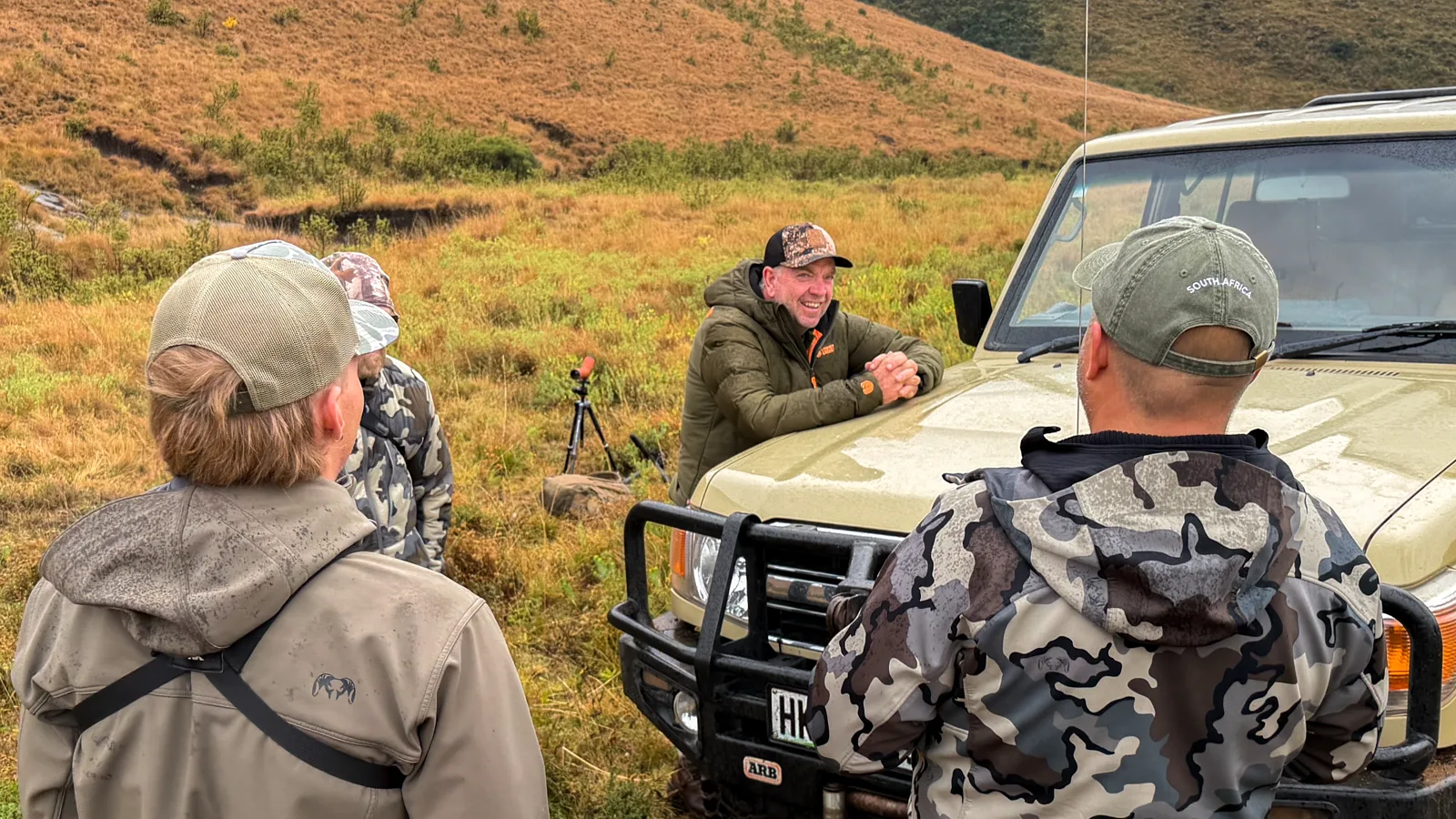 Safari group wearing layered weather-resistant clothing beside a hunting vehicle